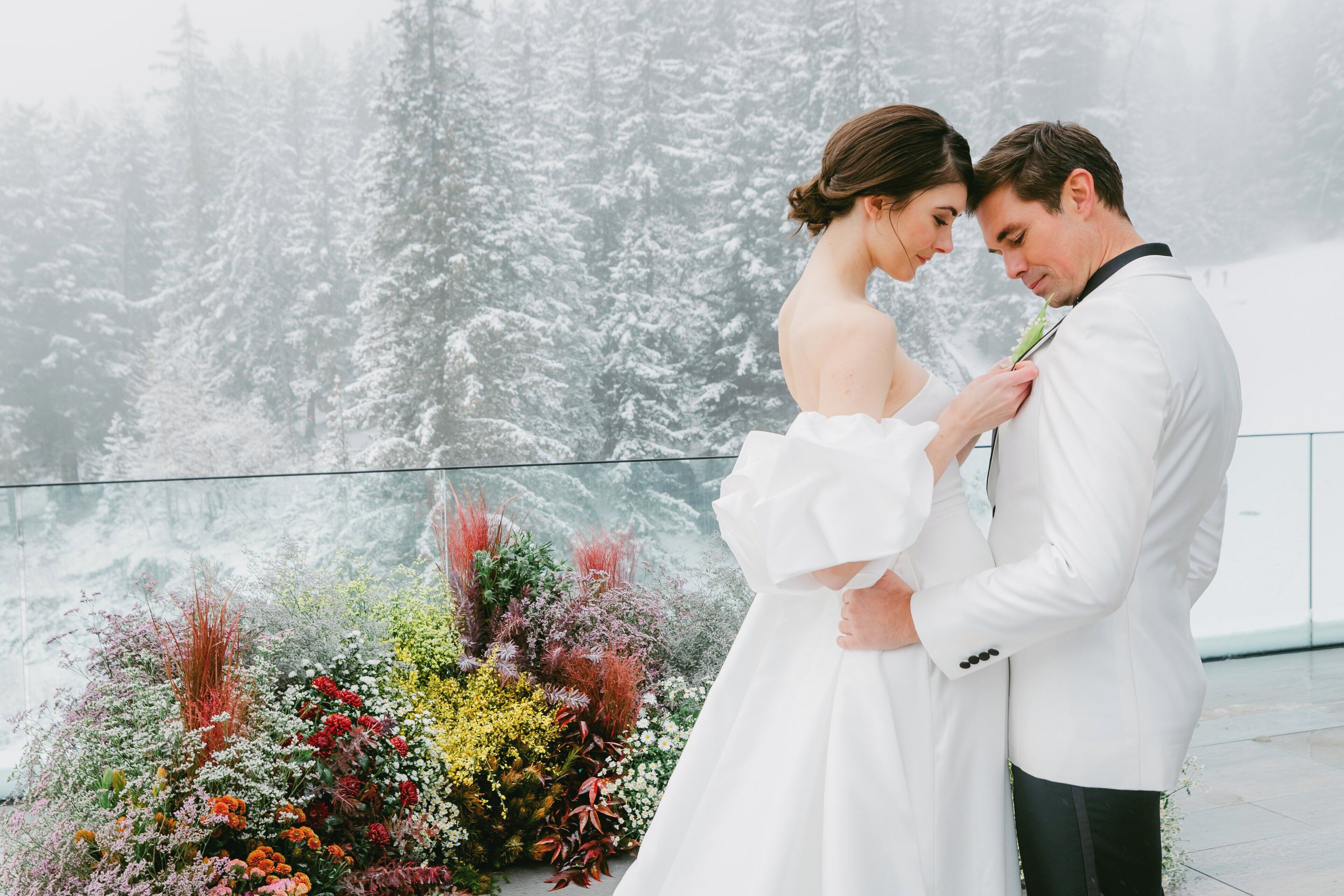 A couple in wedding attire standing in front of a floral arrangement with a snowy forest backdrop.
