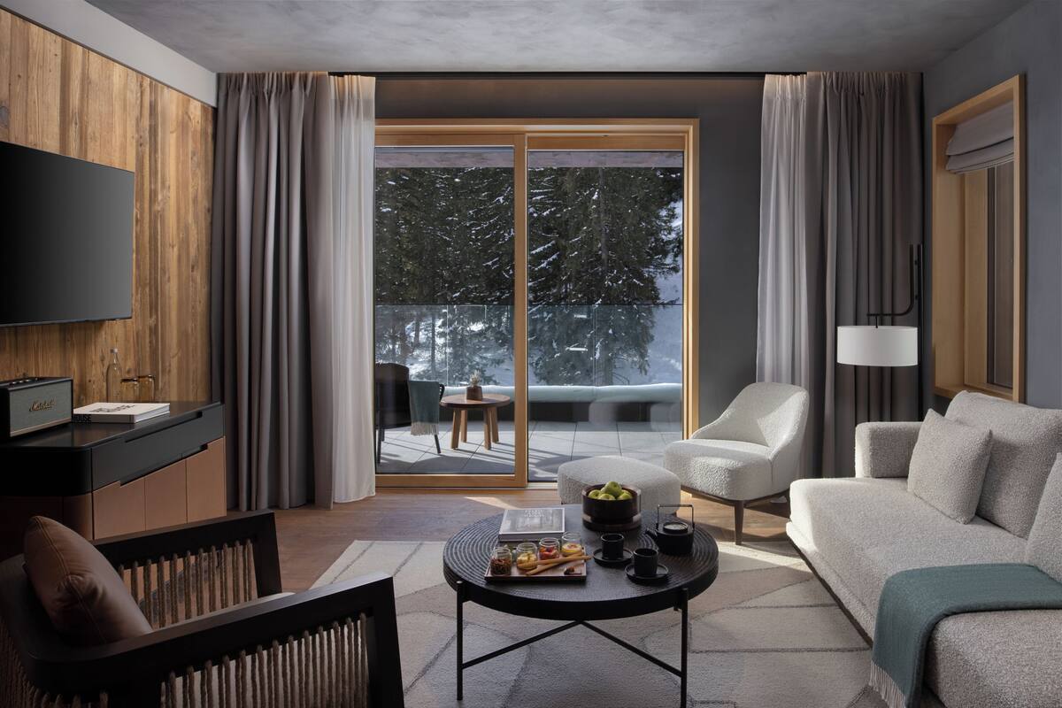 A modern living room with a sofa, armchair, and coffee table. A flat-screen TV is mounted on the wall, and there is a sliding glass door leading to a balcony.