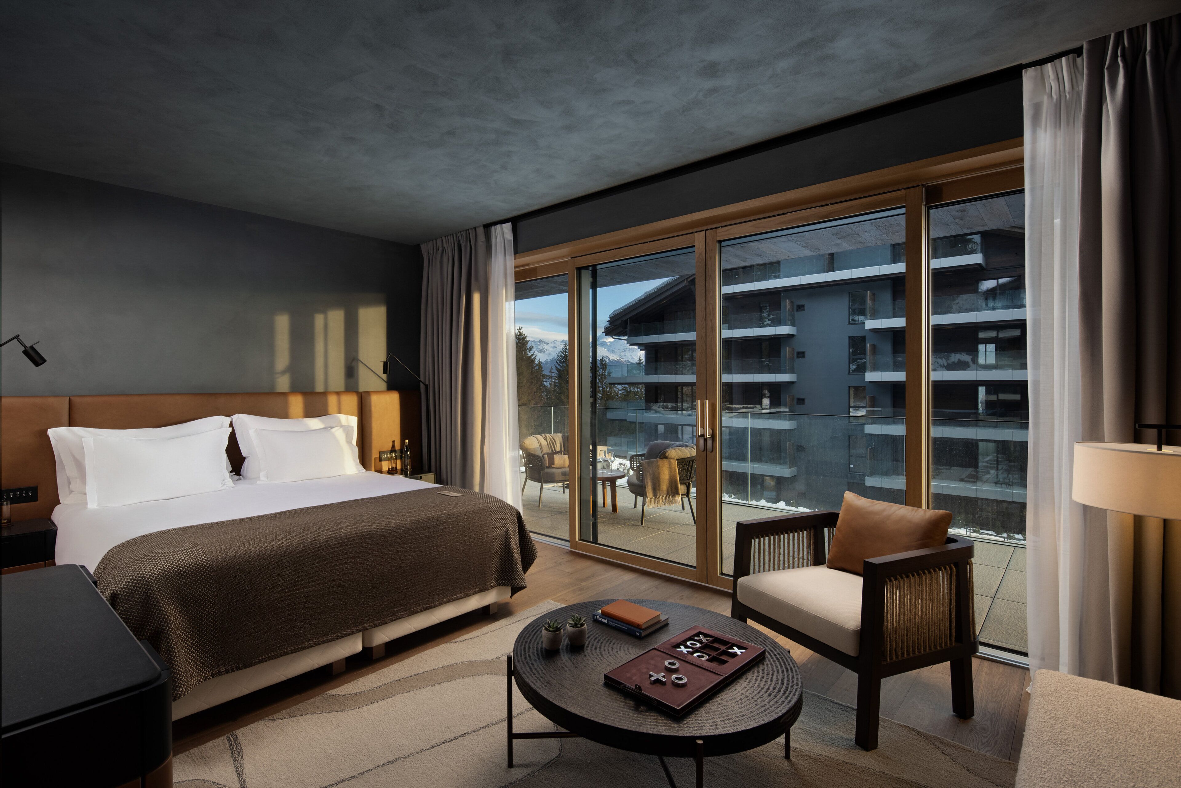 A hotel room with a large bed, a seating area with a table, and a view of a building through a glass door.