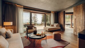 Una sala moderna con sofá, sillón y reposapiés, una mesa de centro redonda con aperitivos, y vistas a un comedor y árboles a través de amplias ventanas.