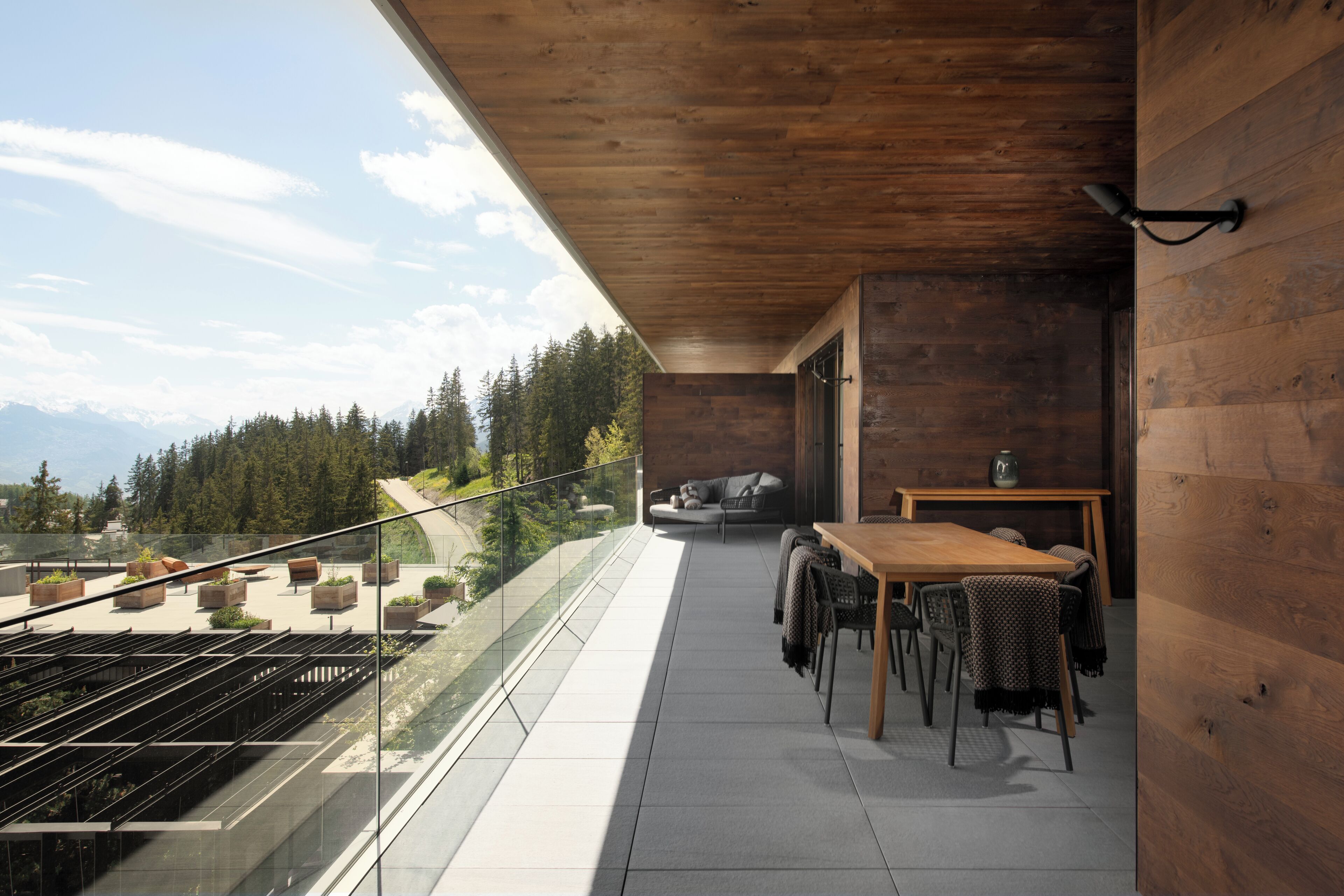 Un balcon avec une table et des chaises en bois, un canapé et une vue sur une forêt et des montagnes.