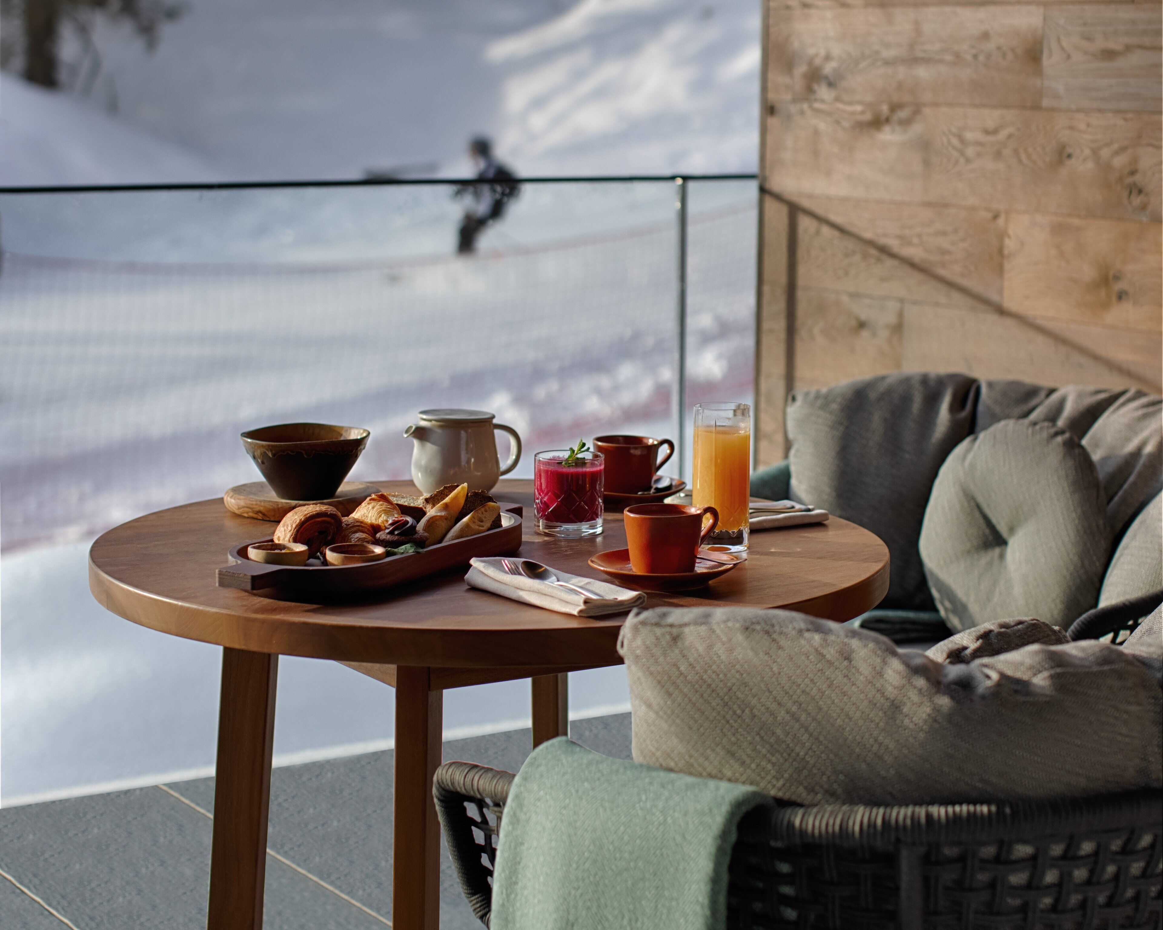 A breakfast table with pastries, a jug, and a glass of orange juice on a balcony with a snowy view.