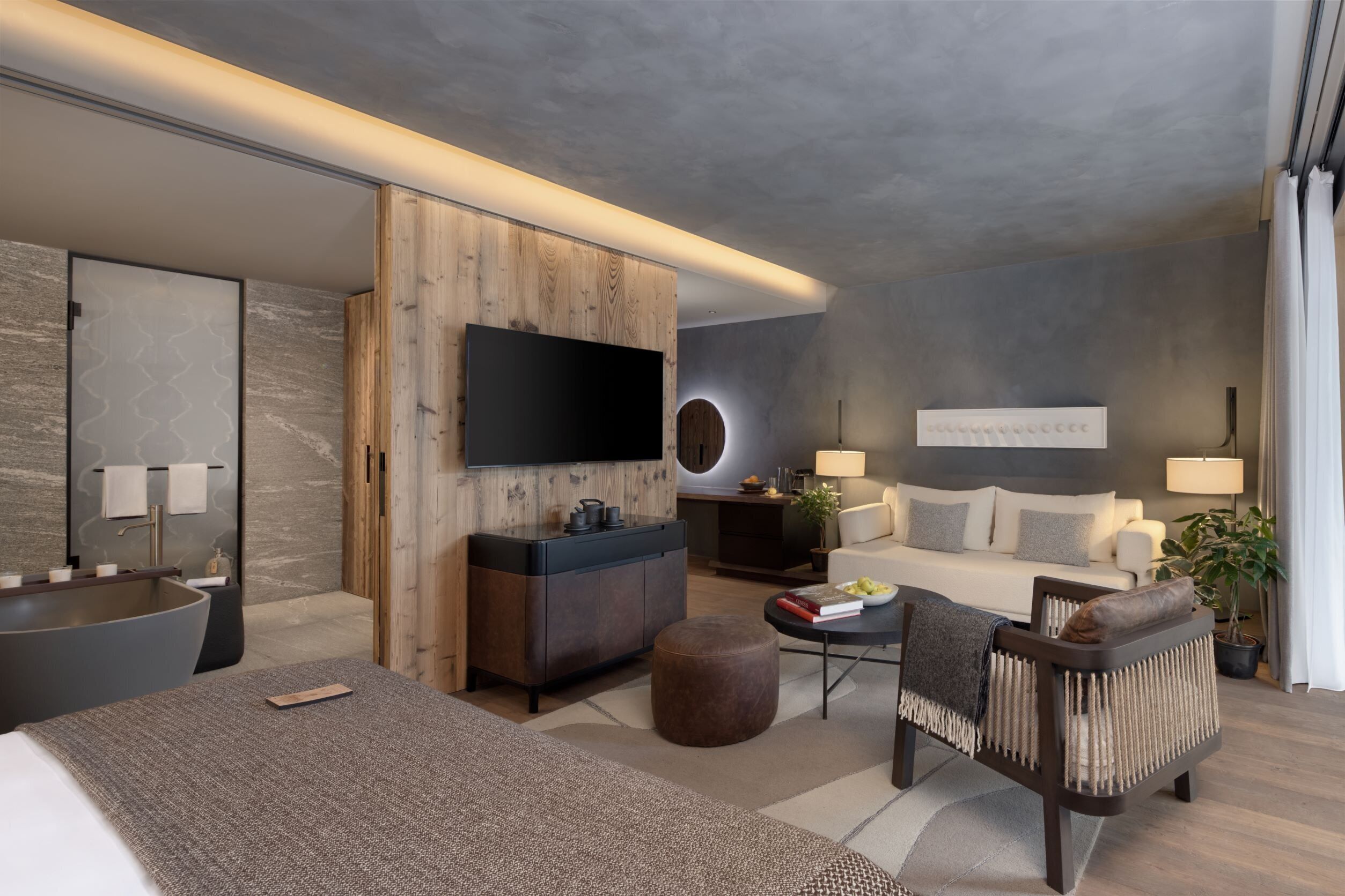 Habitació d'hotel moderna amb un llit gran, zona de seients amb un sofà i una butaca, taula petita amb llibres i una làmpada, televisió penjada a la paret i un moble de fusta.