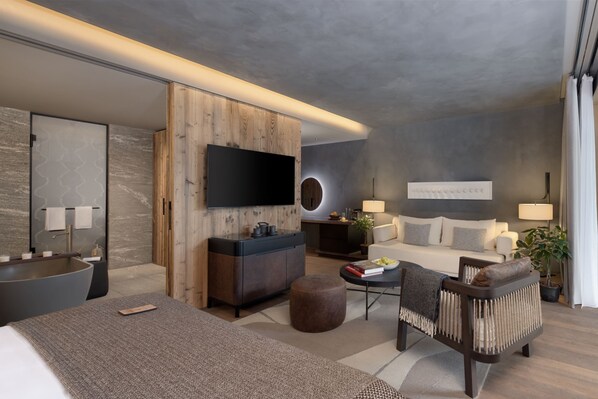 Una habitación de hotel moderna con una cama grande, una zona de estar con sofá y sillón, una mesita con libros y una lámpara, un televisor montado en la pared y un armario de madera.
