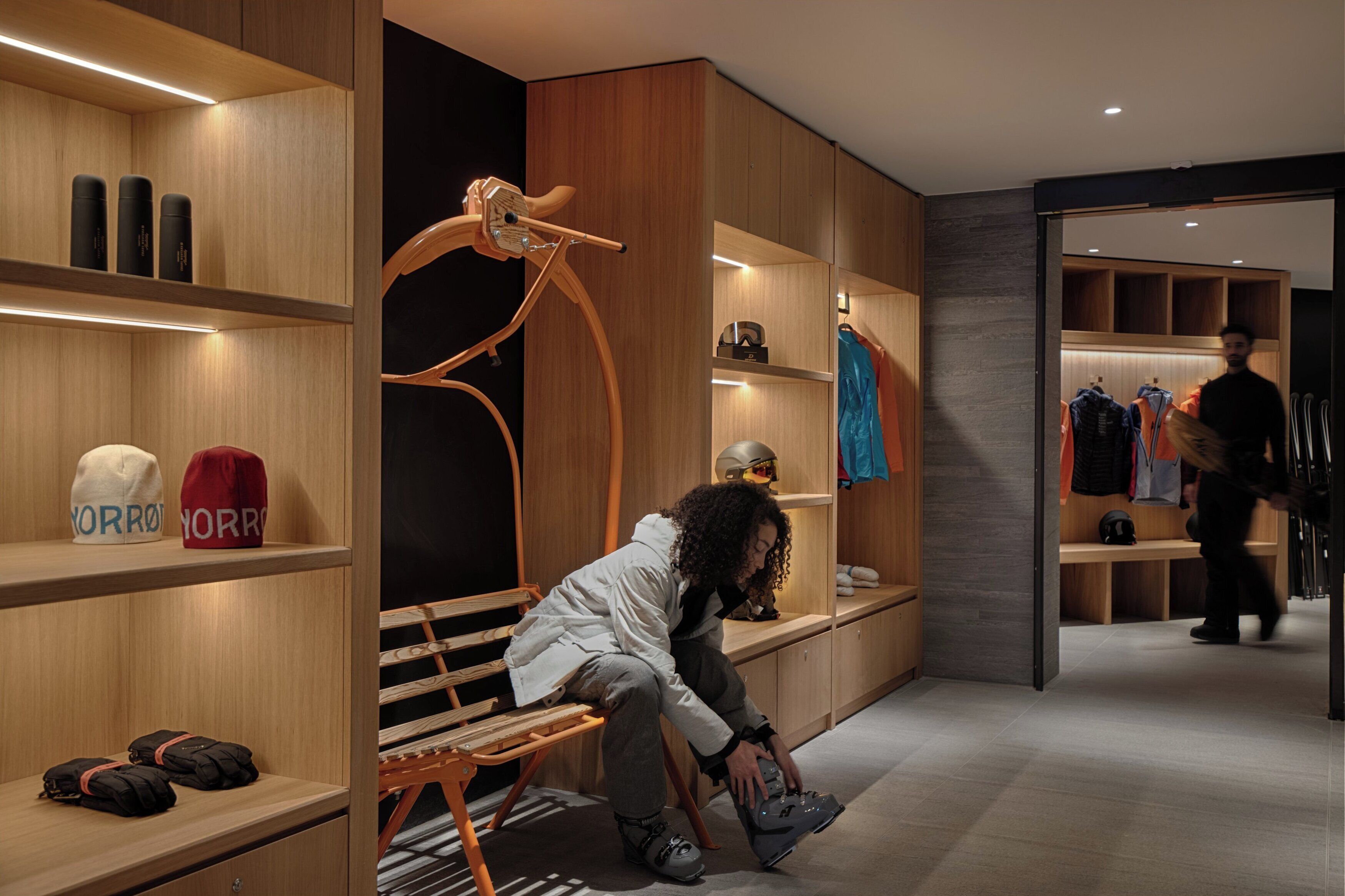 Interior moderno de hotel con estanterías de madera, un banco y una persona ajustándose los zapatos.