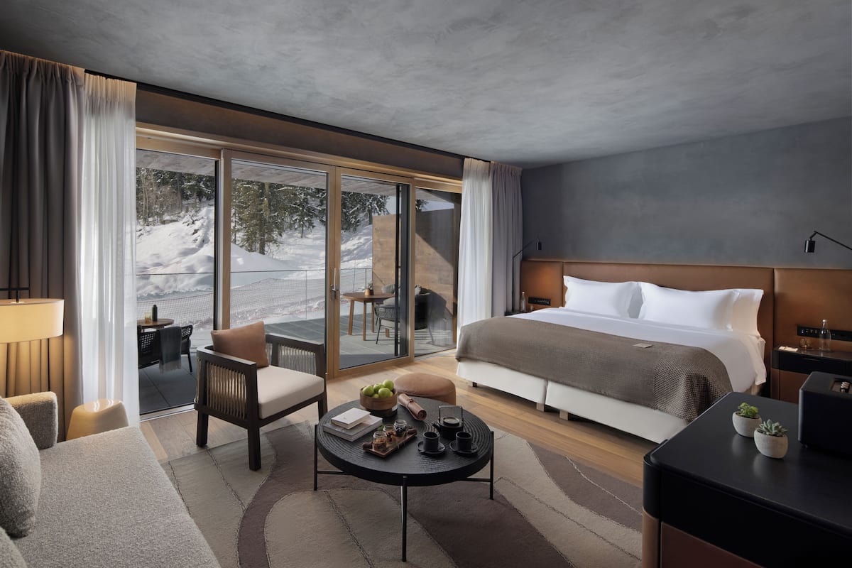 A modern hotel room with a large bed, a dining area with a table and chairs, and a view of a snowy landscape through large windows.
