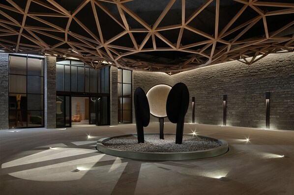 Un interior moderno con una estructura de techo geométrica, una zona de estar circular central y una pared de piedra.
