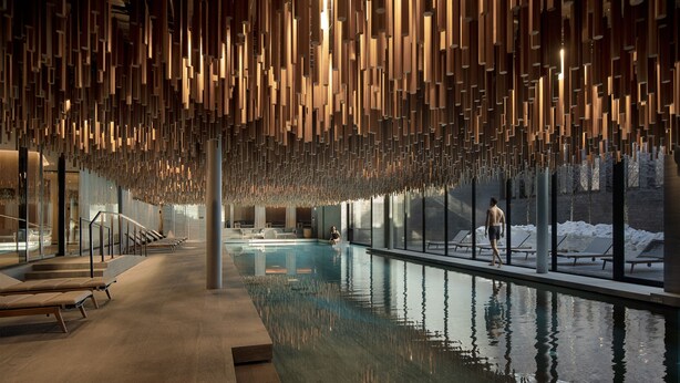 A modern indoor pool area with a reflective water surface, wooden ceiling panels, and a view of a snowy landscape through large windows. - Six Senses Crans-Montana (Lens)