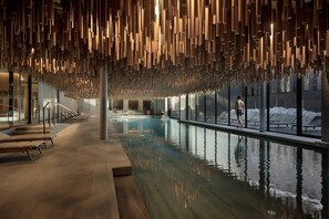 Un área de piscina interior moderna con una superficie acuática reflectante, paneles de techo de madera y vistas a un paisaje nevado a través de amplios ventanales.