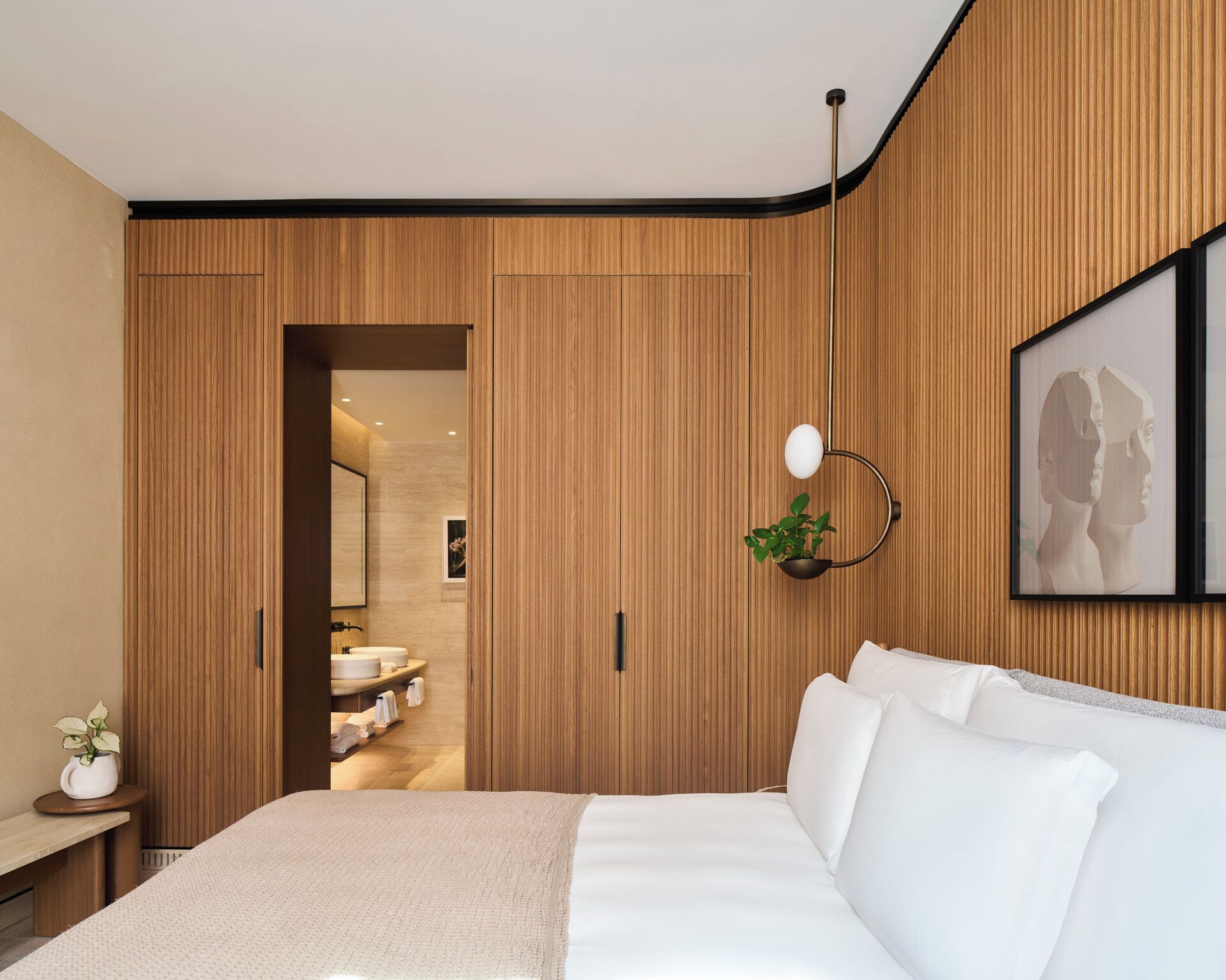 Un dormitorio moderno con pared de paneles de madera, una cama con ropa de cama blanca, una mesita de noche con una planta y una obra de arte enmarcada en la pared.