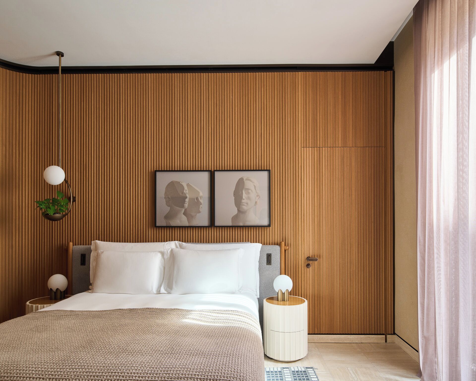 A bedroom with a wooden panel wall, a bed with white linens, two framed pictures, a bedside lamp, and a small round table with a plant.