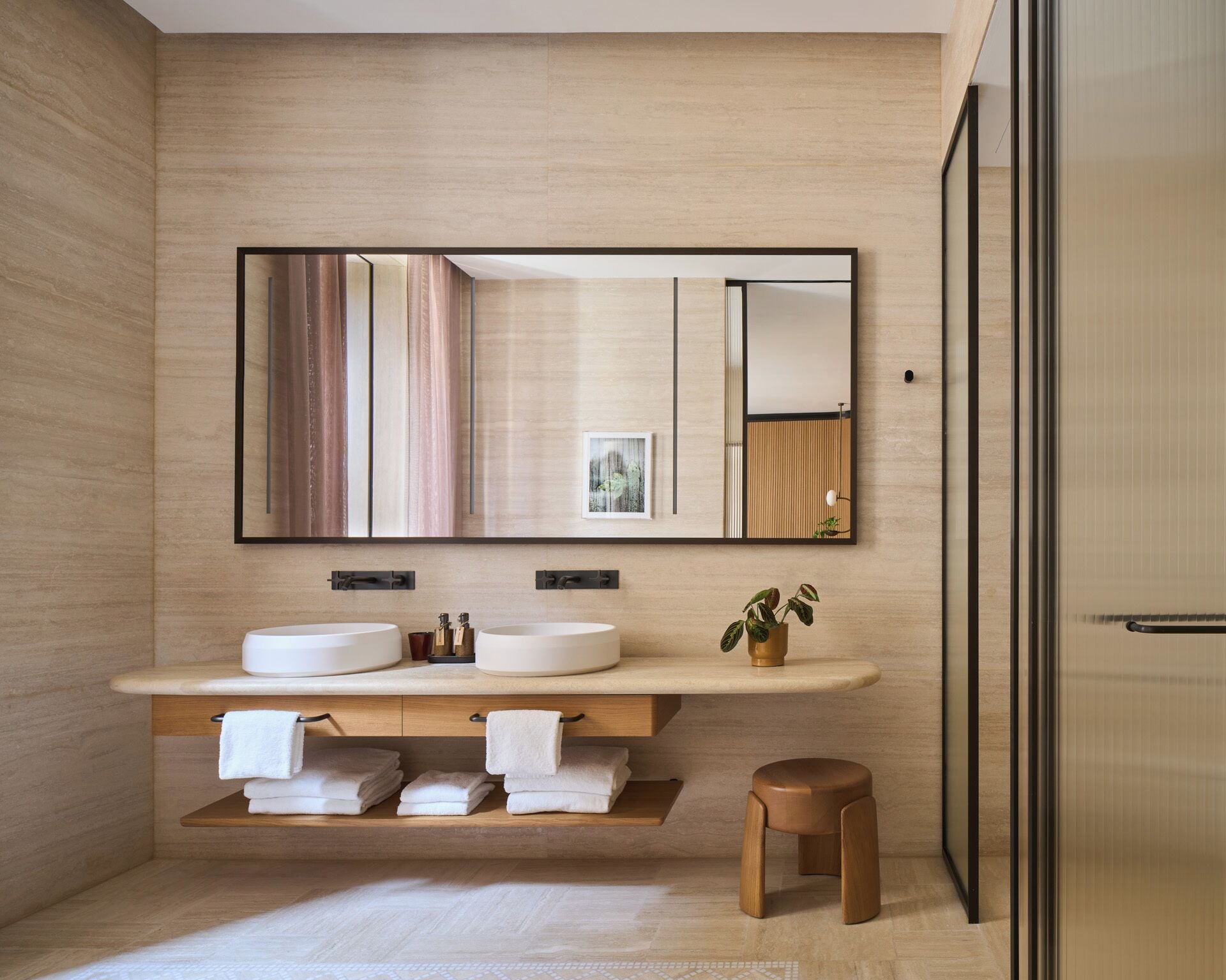 A modern bathroom with a wooden vanity, two sinks, a large mirror, and a small stool.