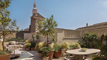An outdoor patio with a round table and chairs, surrounded by potted plants and a view of a building with a tower.