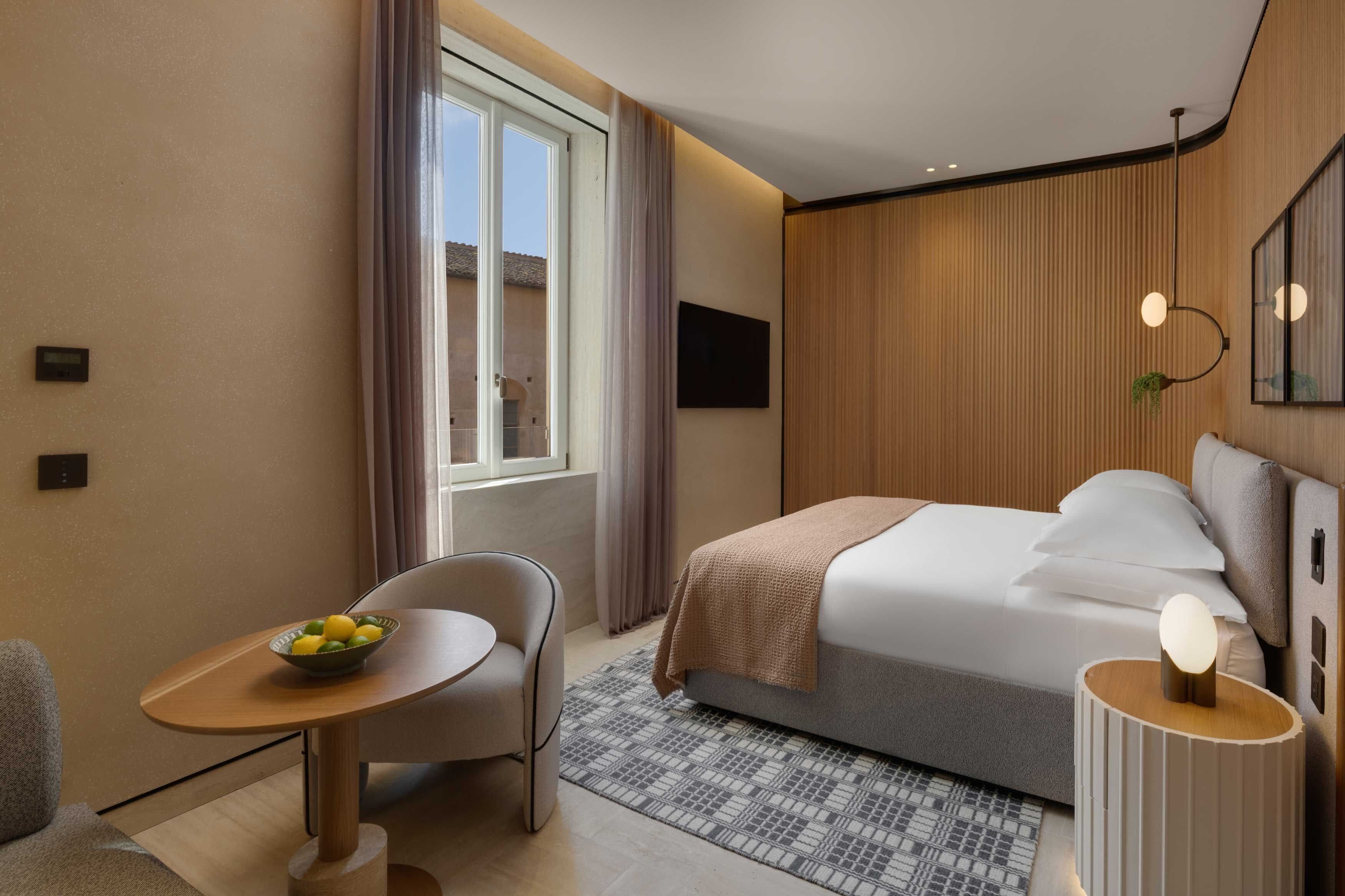 Habitación de hotel con cama, escritorio con frutero, silla, mesita de noche con lámpara y ventana con cortinas.