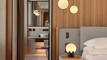 A modern hotel room with a wooden panel wall, a round bedside table with a lamp, a bed with white linens, and a view into the bathroom.