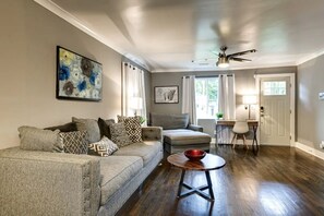 Ein Wohnzimmer mit einer Couch, einem Sessel und einem kleinen Tisch mit einer Schale. Es gibt einen Deckenventilator, ein Gemälde und ein Fenster mit Vorhängen.