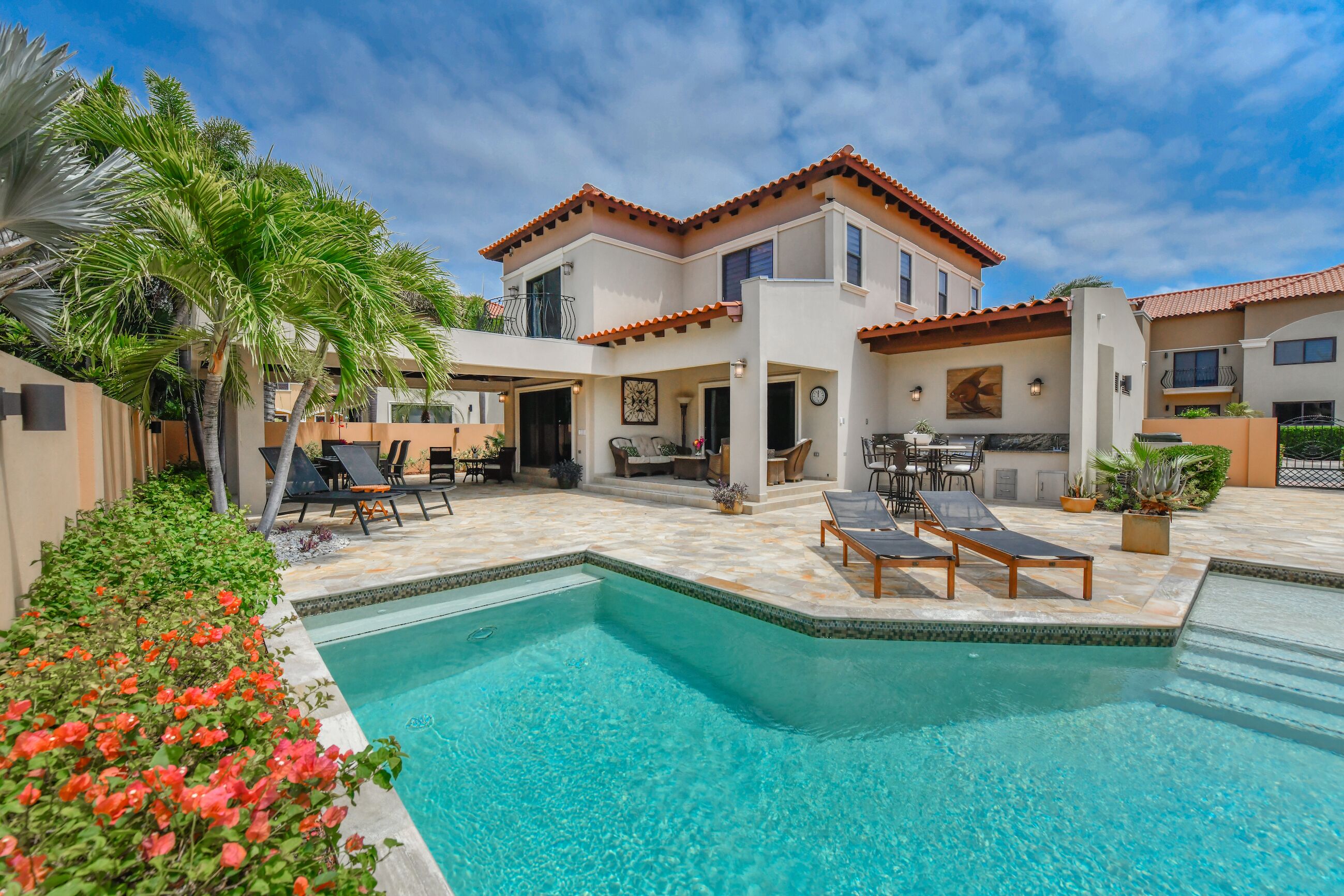 Une maison à deux étages avec une piscine, du mobilier d’extérieur et un jardin agrémenté de palmiers et de fleurs.