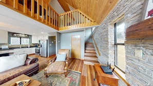 A spacious living area with a high ceiling, a wooden staircase, a stone wall, and a kitchen with modern appliances.