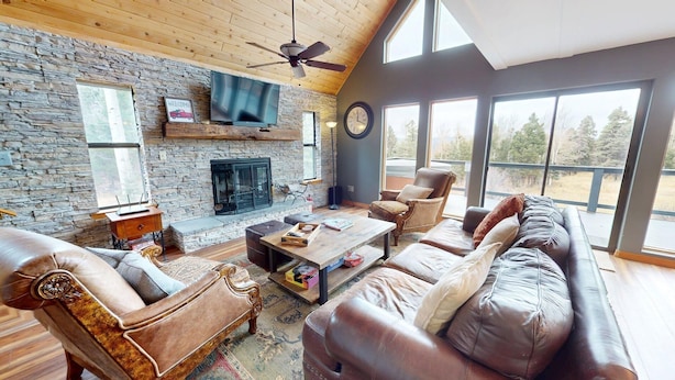 A cozy living room with a stone fireplace, leather sofas, and a large window offering a view of the outdoors.