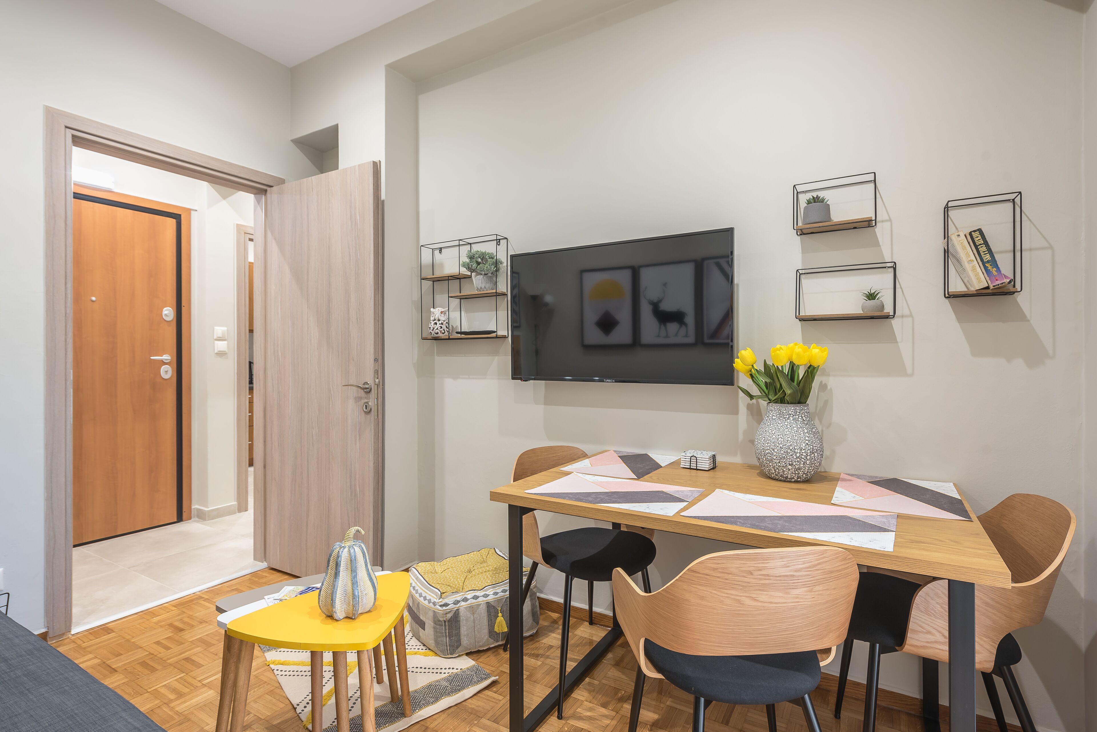 A dining area with a wooden table, chairs, and a vase of yellow flowers. There are wall-mounted shelves with plants and books.