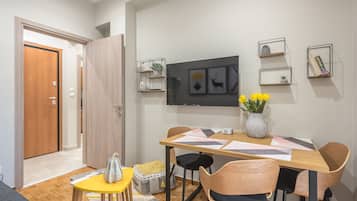 A dining area with a wooden table, chairs, and a vase of yellow flowers. There are wall-mounted shelves with plants and books.