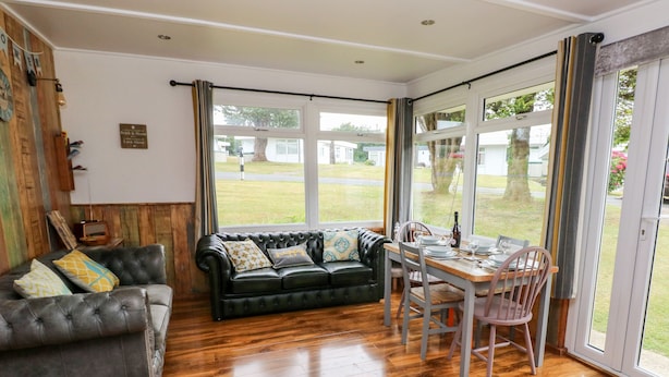 Una sala de estar acogedora con un sofá de cuero, una mesa de comedor preparada para dos personas y ventanas amplias con vistas al exterior.