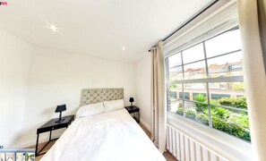 Ein Schlafzimmer mit einem großen Bett, zwei Nachttischen mit Lampen, einem Fenster mit Blick auf Grünflächen und Gebäude sowie einem Heizkörper unter dem Fenster.