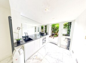 Eine moderne Küche mit schwarzer Arbeitsplatte, weißen Schränken und einem Spülbecken. Linkerhand steht ein Blumentopf und vor der Tür ist ein Grill.