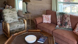 Ein gemütliches Zimmer mit Holzwänden, einem Deckenventilator, einem Sofa mit gemusterten Kissen, einem Holztisch mit einer Schale und einem Buch sowie einem Fenster mit Vorhängen.