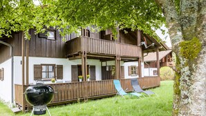 Une maison en bois à deux étages, dotée d’un balcon, d’un barbecue et de deux chaises longues dans la cour.