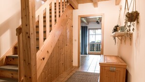 Un escalier en bois mène à une pièce dont le sol est recouvert de carreaux, qui contient un petit meuble et où sont suspendues des plantes.
