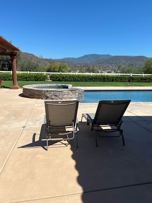 A pool area with two lounge chairs and a stone hot tub, surrounded by a well-maintained lawn and mountains in the background.