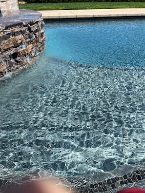 A clear swimming pool with a stone waterfall feature and a stone ledge.