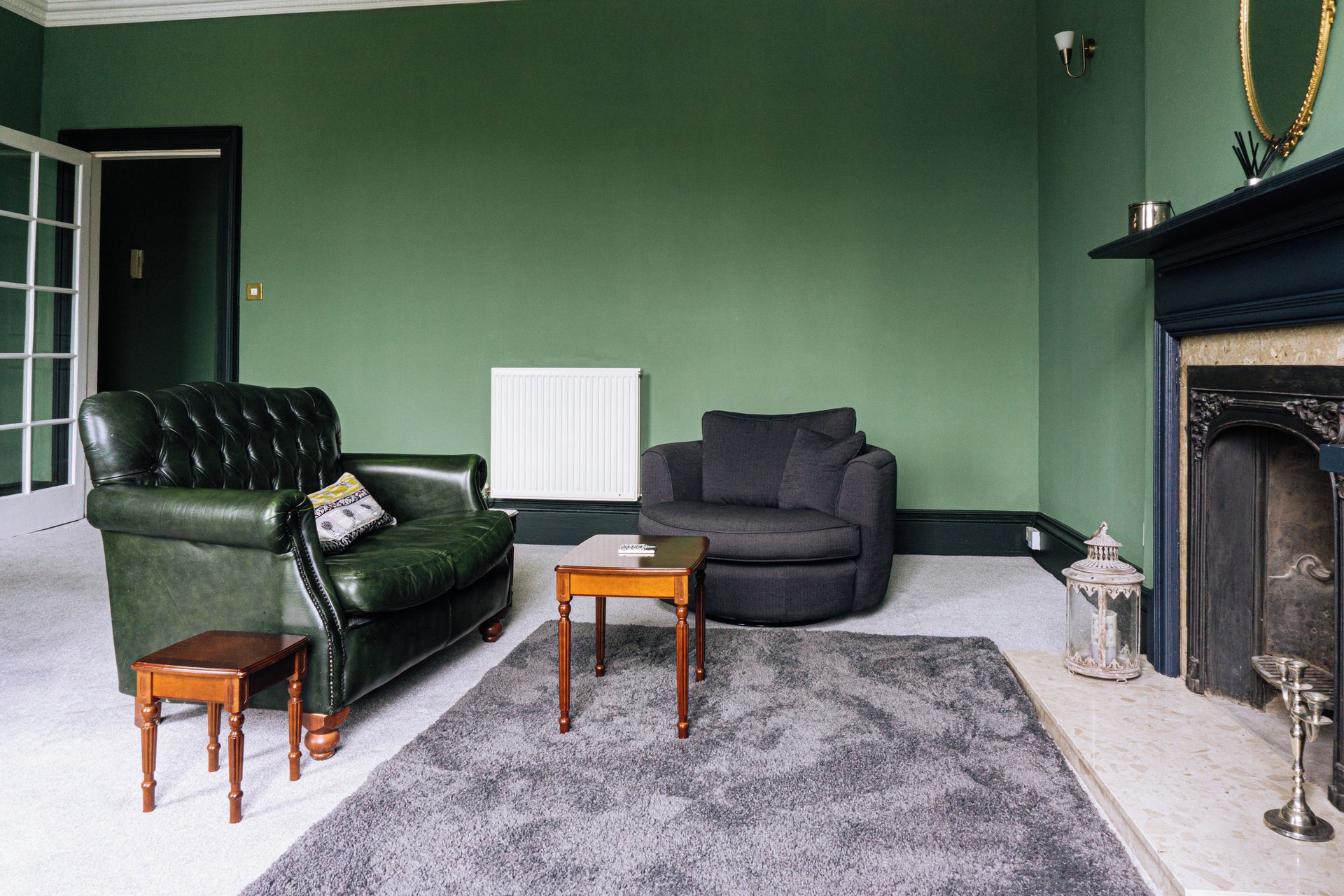 A room with a green wall, a leather sofa, a grey armchair, a small wooden table, and a fireplace.