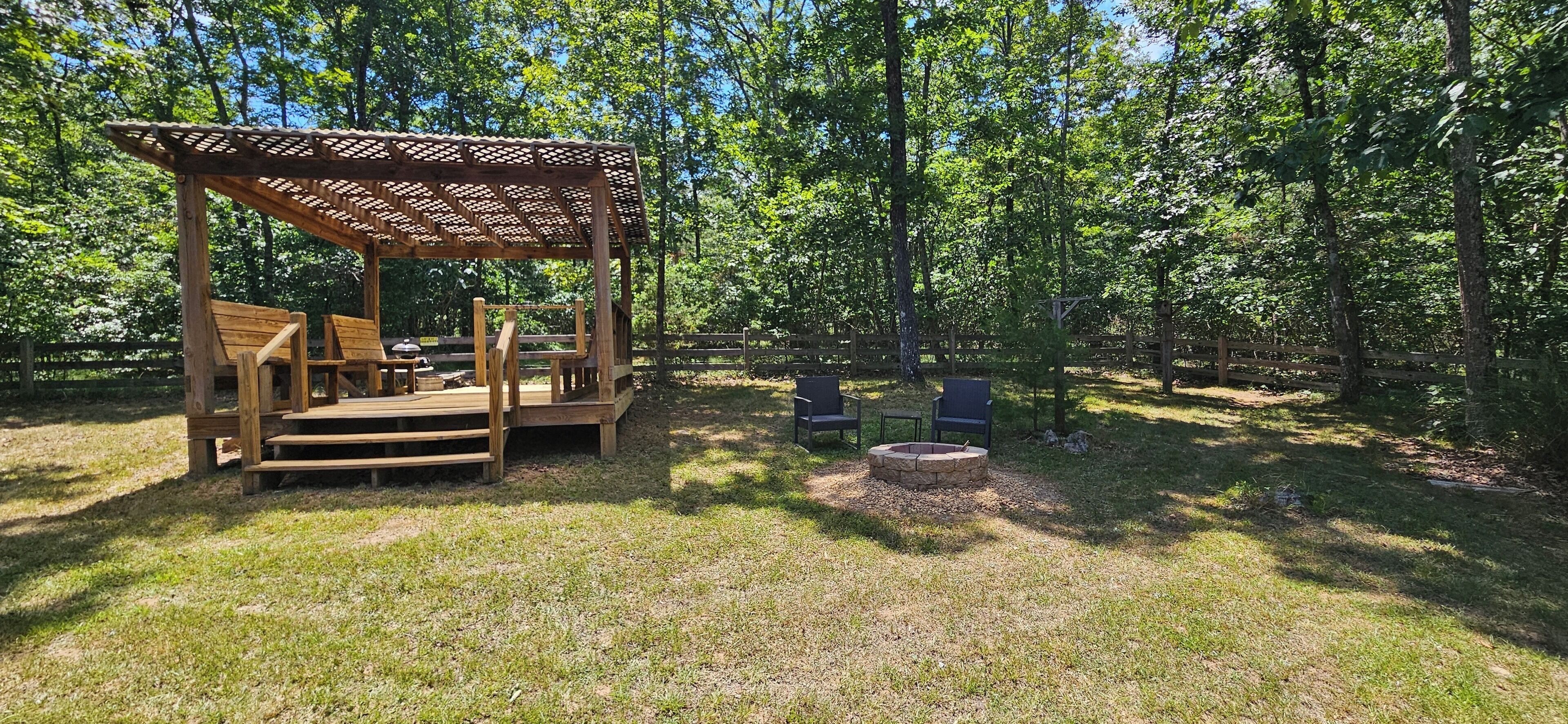 Une terrasse en bois avec des sièges et un foyer, située dans un espace boisé.