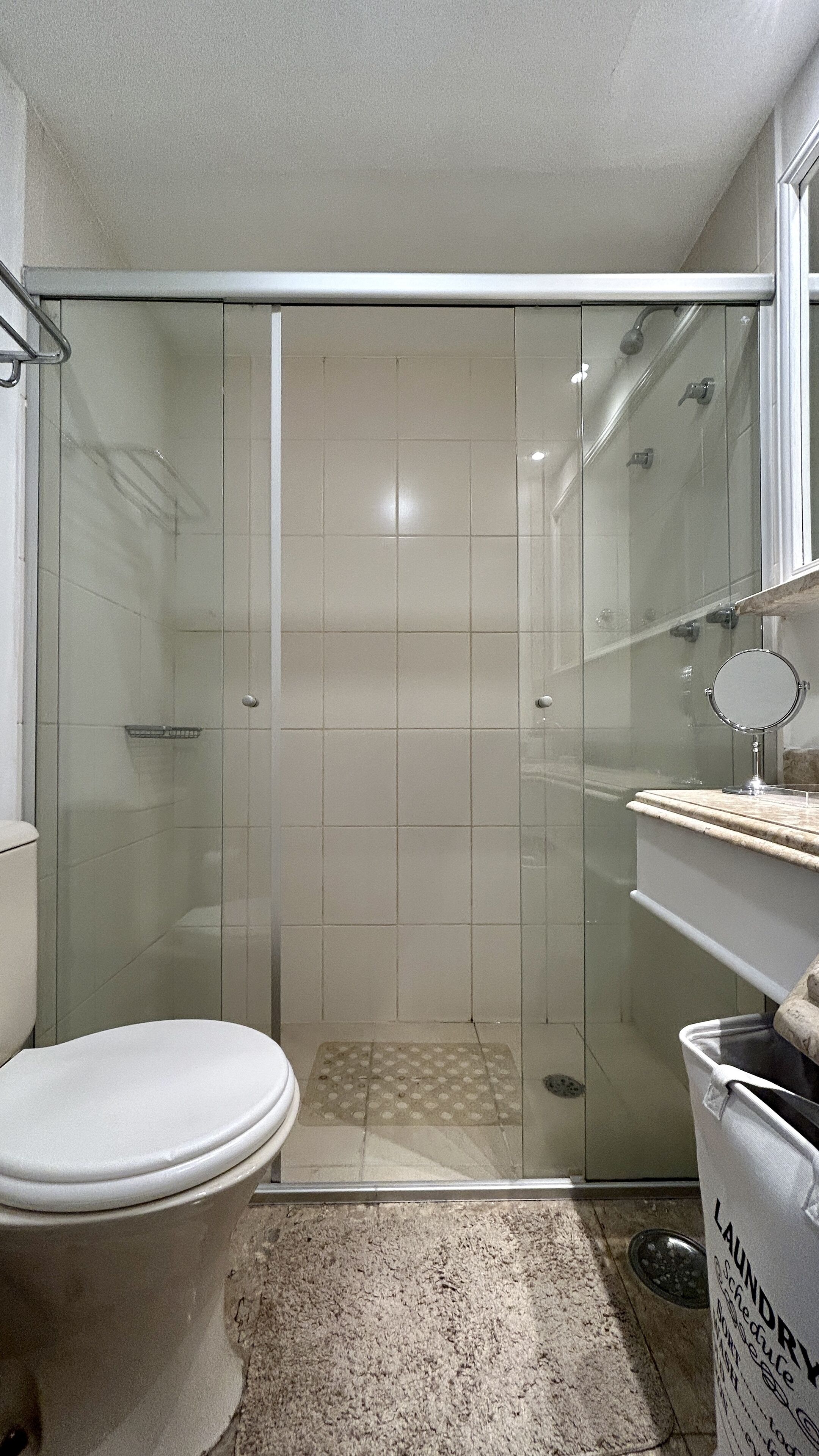 Une salle de bain avec une douche à parois de verre, des toilettes, un lavabo avec un miroir et un panier à linge.