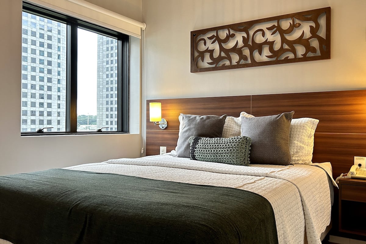 A hotel room with a large bed, two bedside lamps, a decorative wall piece, and a window with a view of buildings.