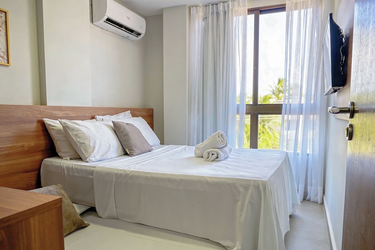 A hotel room with a wooden headboard, a bed with white linens, a rolled towel, and a TV mounted on the wall.