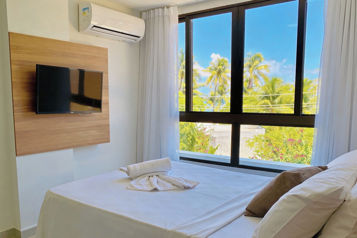 A hotel room with a bed, a TV mounted on the wall, and a window with a view of palm trees and greenery.