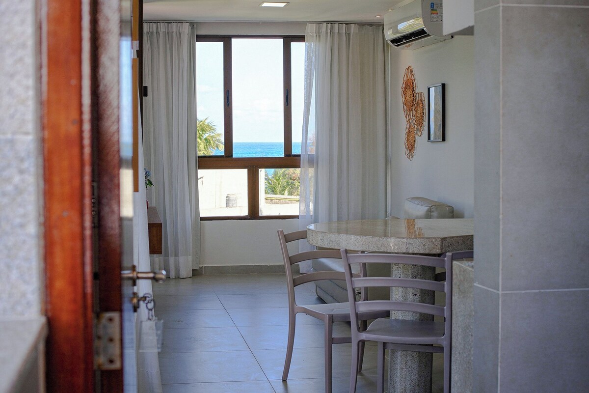 A room with a dining area, a window with a view of the sea, and a wall-mounted air conditioner.