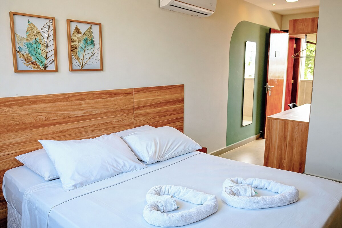A neatly made bed with white linens and two towels, a wooden headboard, and two framed leaf artworks on the wall.