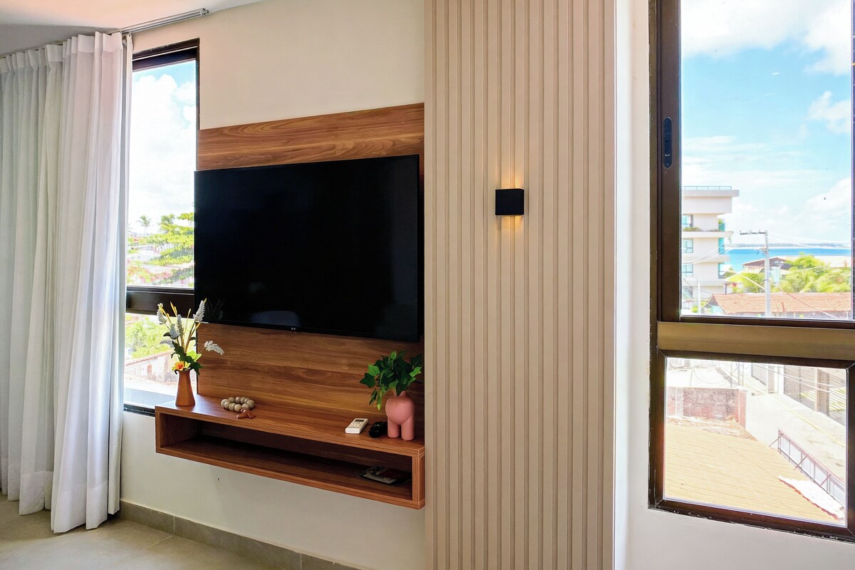A modern living room with a large flat-screen TV mounted on a wooden panel, a small shelf with decorative items, and a view of a coastal area through a large window.