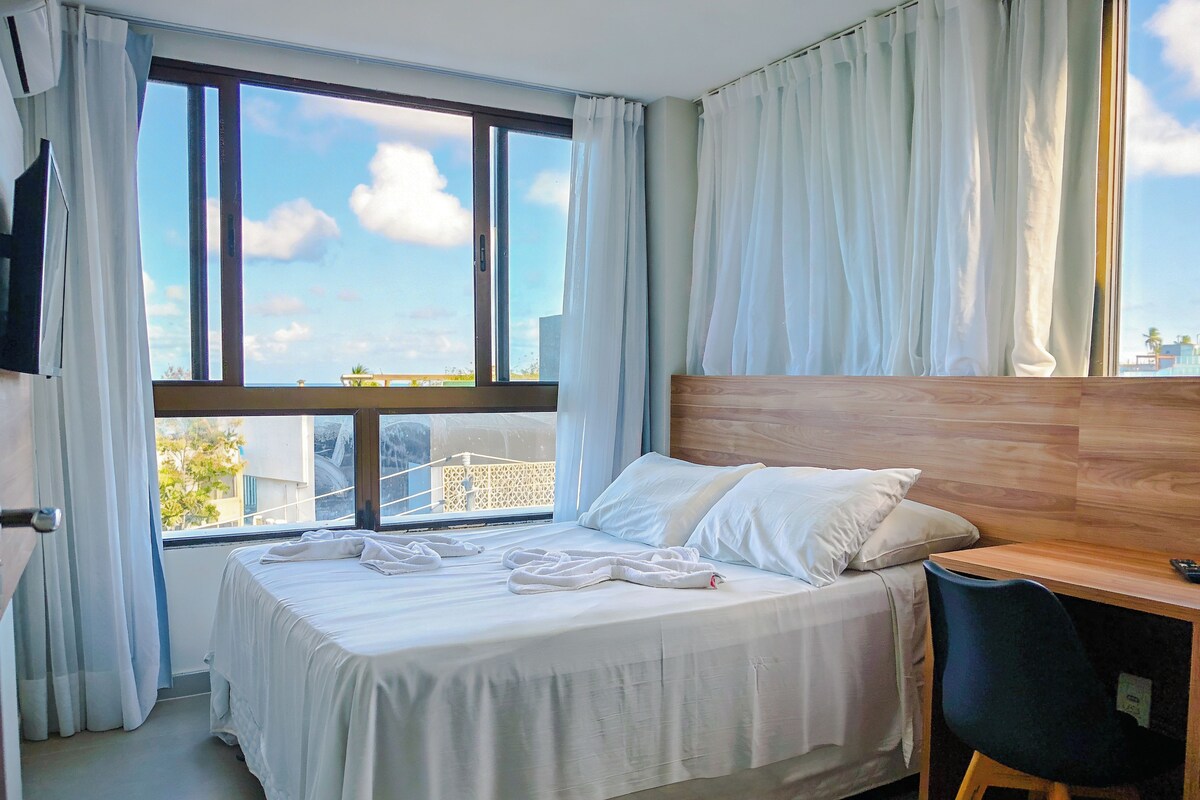 A hotel room with a bed, a desk, and a view of the cityscape through large windows.