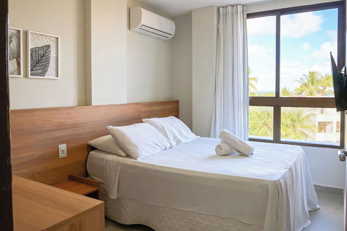 A hotel room with a bed, a wooden nightstand, a wall-mounted air conditioner, and a window with a view of palm trees.