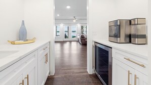Una cocina moderna con armarios blancos, detalles dorados y electrodomésticos de acero inoxidable. Un florero y una bandeja están sobre la encimera. La cocina da a un comedor con vistas al exterior.