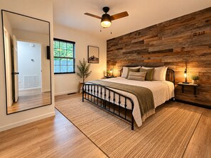 A bedroom with a wooden wall, a bed with a headboard, a ceiling fan, a window with a view, and a door.