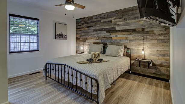A bedroom with a bed, a ceiling fan, a window, and a wall-mounted TV.