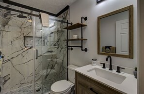 Un baño con ducha de mampara de vidrio, inodoro blanco, lavabo con tocador de madera y espejo con marco de madera.