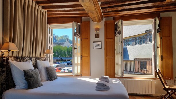 Un dormitorio con una cama grande, vigas de madera, vista a la calle y una silla junto a la ventana.