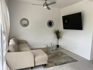 A living room with a beige sofa, a glass coffee table, two decorative mirrors, a ceiling fan, and a flat-screen TV mounted on the wall.